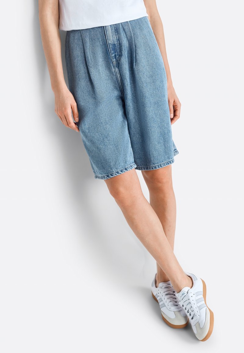 Pantalones cortos de mezclilla en azul claro con un ajuste relajado, que presentan pliegues y un dobladillo deshilachado. Combinados con zapatillas blancas y una blusa blanca casual.