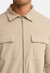 Beige kordfløjljakke med lynlås foran og to brystlommer båret af en person, som viser krave og overkrop.