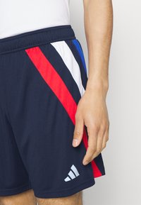 Marina atletiska shorts med diagonala ränder i röd, vit och blå. Inkluderar en vit Adidas-logotyp på nedre vänstra sidan.