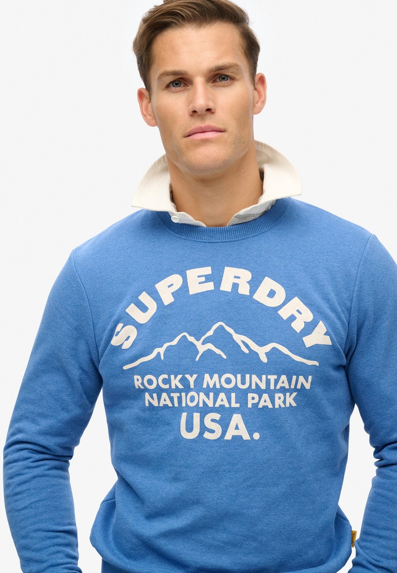 Sudadera azul con cuello redondo, que presenta un gráfico de montañas blancas y el texto "SUPERDRY ROCKY MOUNTAIN NATIONAL PARK USA." en la parte frontal.