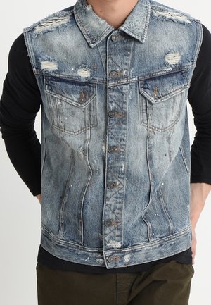 Verwaschene blaue, ausgewaschene Jeansweste mit Knopfverschluss, Brusttaschen, getragen über einem schwarzen Langarmshirt.