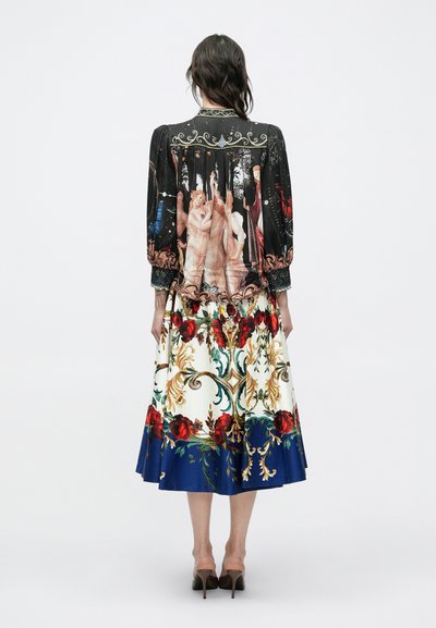 Frau trägt eine schwarze Bluse mit einem klassischen Gemäldeaufdruck und einen weißen, reich verzierten Rock mit roten Rosen und blauem Saum, steht mit dem Rücken zur Kamera.