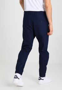 Marineblauwe tapered broek met elastische boorden, achterzakken en een gladde textuur, gecombineerd met witte sneakers en zichtbare sokken.