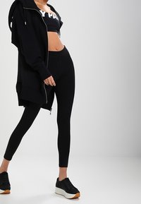Black oversized hoodie, cropped tank top with white text, form-fitting black leggings, and black sneakers with a white sole and brown accents.