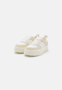 Sneaker beige e bianche con una suola spessa e texture, punta rotonda, lacci davanti e pannelli laterali con dettagli in pelle liscia e tessuto.