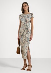 Vestido midi floral com mangas curtas, detalhe de nó na cintura, racha lateral e um fundo bege claro. Usado com sandálias de tacão castanhas e uma bolsa.