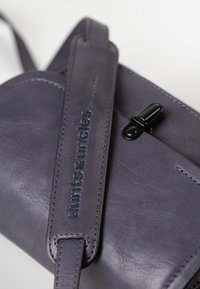Piccola borsa in pelle viola con superficie textured, nome del marchio impresso e una finitura nera della cerniera. I dettagli includono una tracolla cucita.