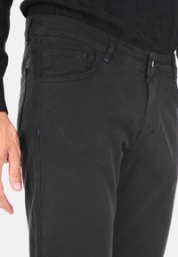 Uomo con pantaloni neri e un maglione nero, con la mano sinistra rilassata accanto ai fianchi su uno sfondo bianco.