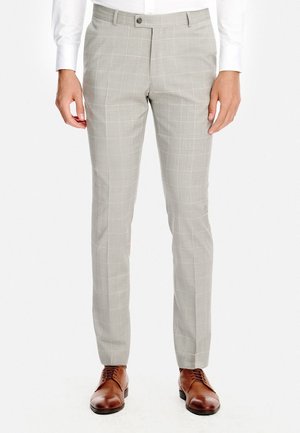 Mężczyzna ubrany w jasnoszare, wąskie spodnie w kratę, brązowe skórzane eleganckie buty i białą koszulę z długim rękawem, stojący na tle białego tła.