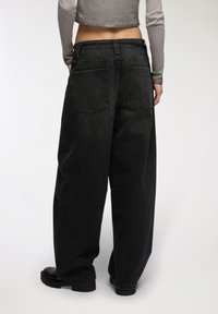 Osoba w luźnych czarnych dżinsach i czarnych butach, z rękami w kieszeniach, w krótkim szarym, długim rękawie, pokazywana tyłem na białym tle.