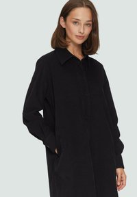 Schwarzes Hemdkleid aus strukturiertem Stoff. Mit kragenförmigem Ausschnitt, Knopfleiste und langen Ärmeln. Seitentaschen sichtbar.
