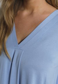 Světle modrý top vyrobený z měkké tkaniny s výstřihem do V a skládaným detailem poblíž výstřihu. Hladká textura bez vzorů.