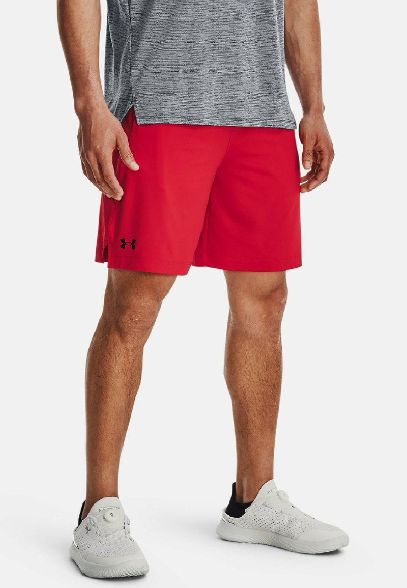 Homme portant un short de sport rouge, un t-shirt gris texturé et des baskets blanches, debout devant un fond blanc.
