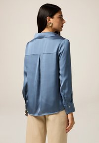Blouse en satin bleu clair avec un col structuré, des manches longues et un détail de pli à l'arrière. Associée à un pantalon beige.