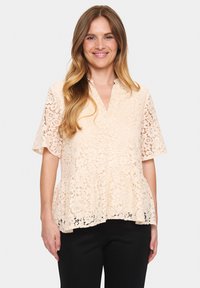 Blusa in pizzo color pesca chiaro con maniche corte e scollo a V. Presenta motivi floreali e un design svasato. Abbinata a pantaloni neri.