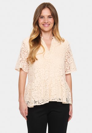 Kanten blouse in licht perzik met korte mouwen en een V-hals. Het heeft bloemmotieven en een uitlopende vorm. Gecombineerd met zwarte broeken.