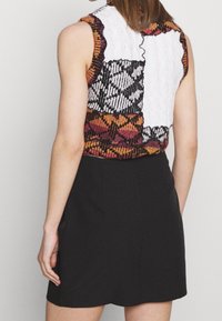 Huoleton toppi, jossa on monivärisiä neulottuja kankaita, valkoinen osa, monimutkaisia kuvioita oranssina ja mustana sekä aaltoileva pääntie.