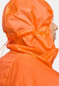 Ljusorange regnjacka med dragskohuva, tillverkad av lättviktsmaterial. Har en slät yta och synliga sömnadsdetaljer.