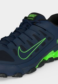 Μπλε ναυτικό αθλητικό παπούτσι Nike με πράσινο λογότυπο Swoosh, πράσινες λεπτομέρειες στη σόλα και μαύρη εξωτερική σόλα, που δείχνει κοντινές λεπτομέρειες από την πλευρά και πάνω.