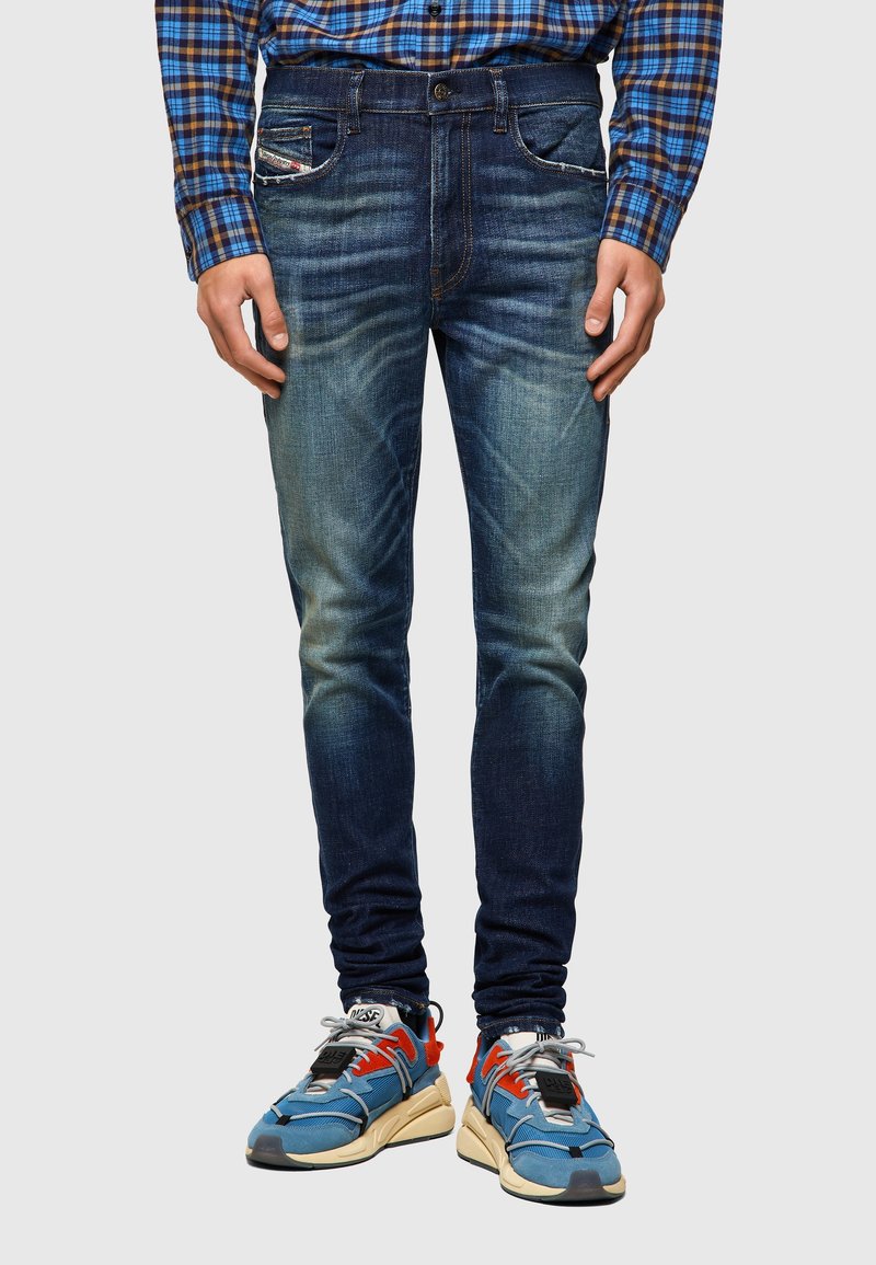 Jean en denim bleu coupe slim porté avec des baskets bleues, orange et beige et une chemise à carreaux bleue et marron, debout devant un fond uni.