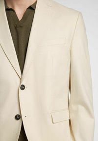 Beżowa marynarka o gładkiej fakturze i klapach z wycięciem, z dwoma czarnymi guzikami i jedną kieszenią na piersi; noszona na ciemnozielonym swetrze.