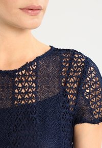 Tummansininen pitsipaita, jossa on läpinäkyvä rakenne. Paidassa on geometrisia kuvioita ja lyhyet hihat, joissa on monimutkaisia kuvioita hartioilla.