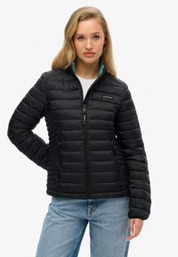 Zwarte gewatteerde jas met horizontale stiksels, ritssluiting aan de voorkant en een staande kraag. Heeft een teal voering en zijzakken. G gedragen met blauwe jeans.