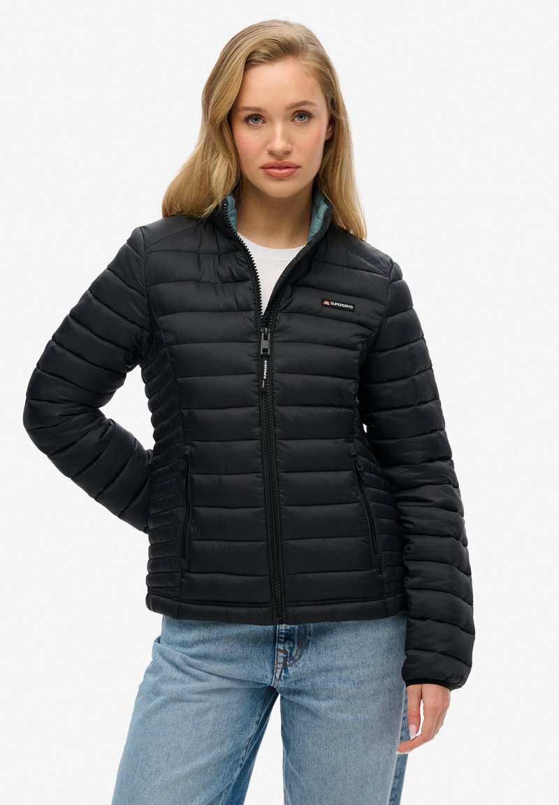 Zwarte gewatteerde jas met horizontale stiksels, ritssluiting aan de voorkant en een staande kraag. Heeft een teal voering en zijzakken. G gedragen met blauwe jeans.