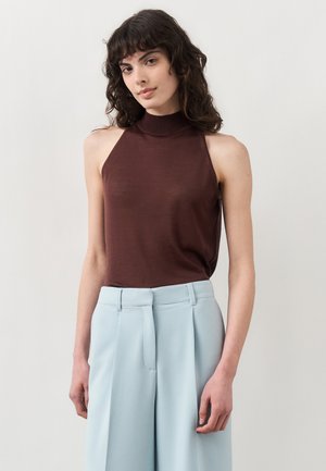 Žena s kučeravými tmavými vlasmi nosiaca hnedý tielkový top a svetlomodré široké nohavice s vysokým pásom na jednofarebnom pozadí.