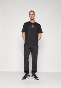 Camiseta atlética negra con un sutil logo gris, combinada con pantalones jogger oscuros y zapatillas deportivas negras con acentos blancos. Textura de fabricación suave.