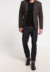 Brun tweedblazer med tre knappar, fickdetalj, kombinerad med en svart crew-neck tröja och mörka denimjeans, avslutad med svarta snörkängor.