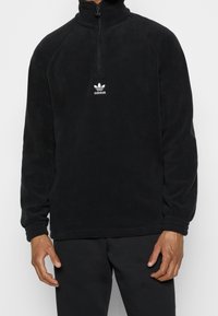 Svart fleece-pullover med hög krage, kvarts dragkedja och ribbade manschetter. Har en liten vit Adidas-trefoliumlogotyp på bröstet.