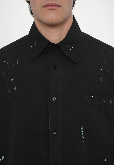 Chemise noire à boutons en coton, avec un col écarté, ornée de motifs irréguliers de éclaboussures de peinture vertes et blanches sur le devant.