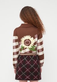 Γυναίκα με σγουρά μαλλιά που φοράει καφέ και κρεμ ζακέτα με καρό σχέδιο και μεγάλο σχέδιο ηλίανθου στην πλάτη, μαζί με σκούρα καρό φούστα.