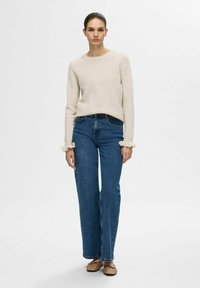Pull en tricot beige à manches longues et bords effilochés, associé à un jean droit bleu taille haute et des ballerines marron clair.