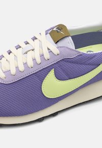 Zapatilla deportiva morada con parte superior de tela texturizada, que presenta un logo de Nike en verde lima, cordones en crema y una suela negra en contraste.