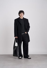 Νεαρός άντρας με σγουρά μαλλιά που φοράει μαύρο παλτό, σκούρο πουλόβερ, φαρδύ μαύρο παντελόνι, μαύρα και άσπρα παπούτσια, κρατώντας μεγάλη μαύρη δερμάτινη τσάντα.