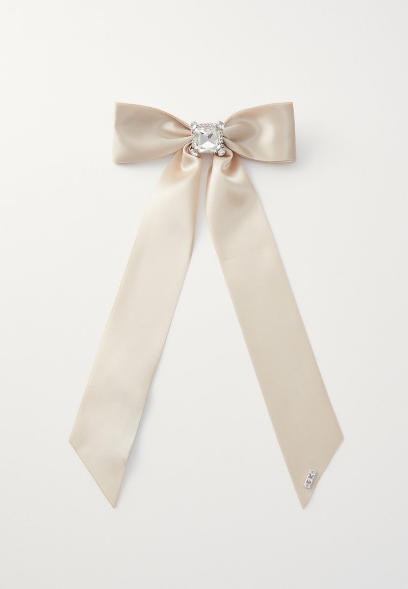 Beige satiinikaari, jossa on kaksi pitkää ja kulmaista häntää sekä keskellä kimalteleva rhinestone-koriste. Sileä pinta, tyylikäs muotoilu.