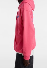 Sudadera rosa con un bolsillo frontal y capucha con cordón. Presenta un logo azul en el pecho. Fabricada con material de algodón suave.
