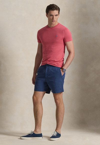 Κόκκινο εφαρμοστό t-shirt με στρογγυλή λαιμόκοψη και μικρό λογότυπο, σε συνδυασμό με μπλε σορτς και ναυτικά sneakers, με casual σχεδιασμό και υφασμάτινη υφή.