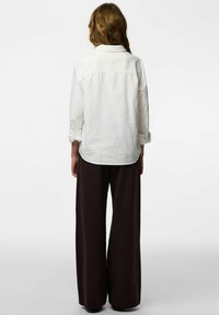 Bijela košulja s dugim rukavima i ovratnikom, s dugmadima na stražnjoj strani te zavrnutim rukavima, uparena s tamnosmeđim širokim hlačama.