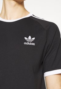 Svart T-shirt med vit ribbad krage och ärmaccenter, med broderad vit Adidas-logotyp ovanför vänster bröst.
