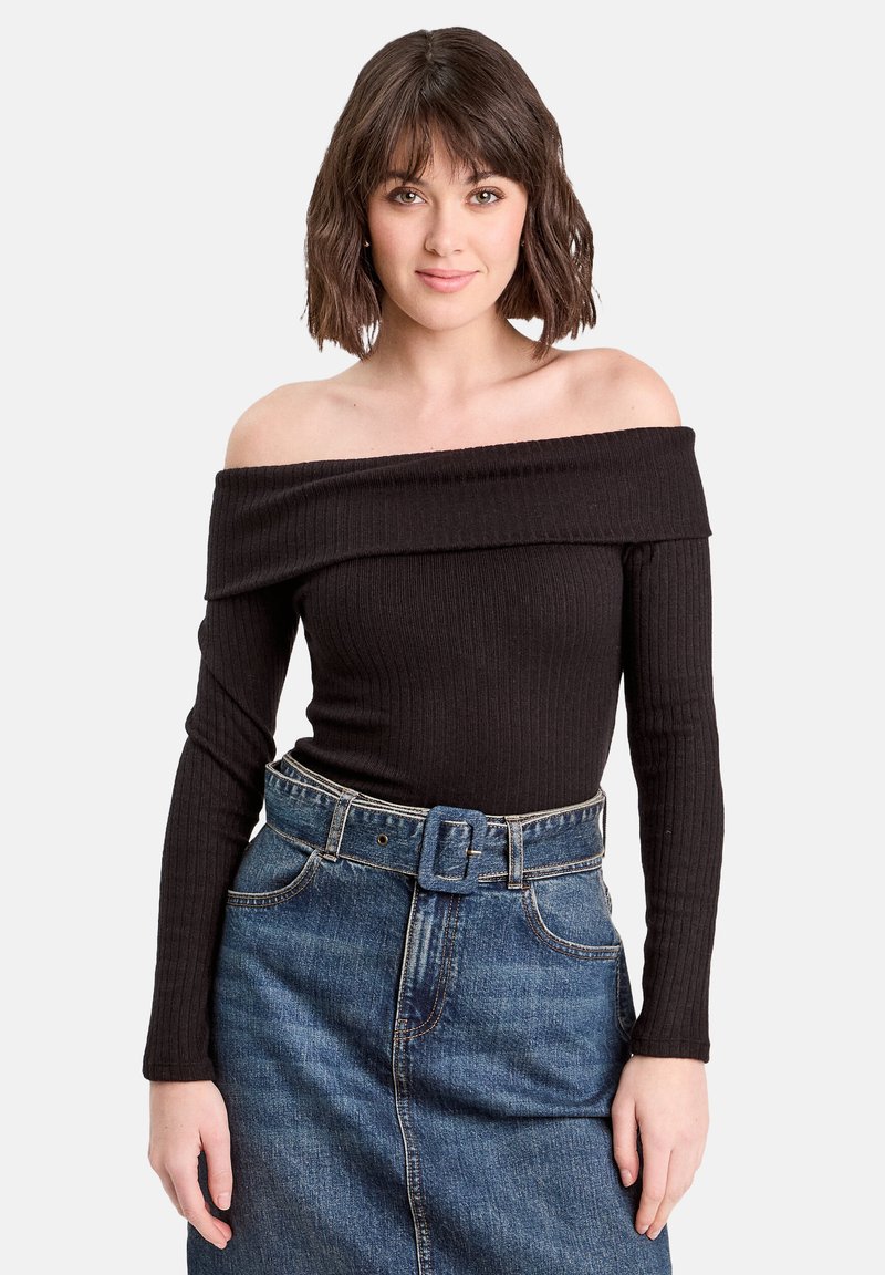 Ung kvinde med kort mørkt hår iført en sort ribbet top med off-shoulder og en højtaljet blå denimnederdel med bælte.
