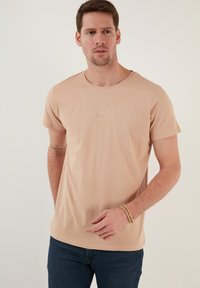 Camiseta de algodón color beige claro con mangas cortas, escote redondeado y detalle de texto en relieve en la parte delantera. Diseño ajustado, textura suave.