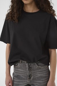 Kobieta w prostym czarnym T-shircie z krótkim rękawem, włożonym do wyblakłych szarych dżinsów, z częściowo widocznymi kręconymi włosami.