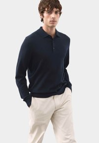 Marineblaues Langarm-Poloshirt aus weichem Stoff, mit Kragen und drei Knöpfen, kombiniert mit hellbeigen Hosen.