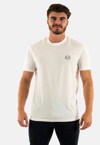 T-shirt bianco in cotone con maniche corte, scollatura rotonda e un piccolo logo circolare nero sul lato sinistro del petto. Design aderente, texture minimal.