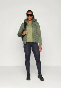 Grøn quiltet vest over en grøn top, kombineret med mørke leggings med paneldetaljer og sorte sko. Modellen bærer solbriller og har en rygsæk med.