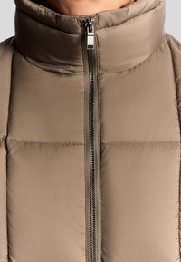 Hellbraune gepolsterte Weste mit hohem Kragen, ausgestattet mit einem zentralen Reißverschluss, glatter Textur und gesteppten Abschnitten für zusätzliche Wärme.