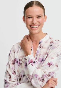 Valkoinen paita, jossa on violetteja kukkakuvioita, pitkät hihat ja rento malli. Pehmeä, tekstuuroitu kangas, jossa on kerätty hihansuu.